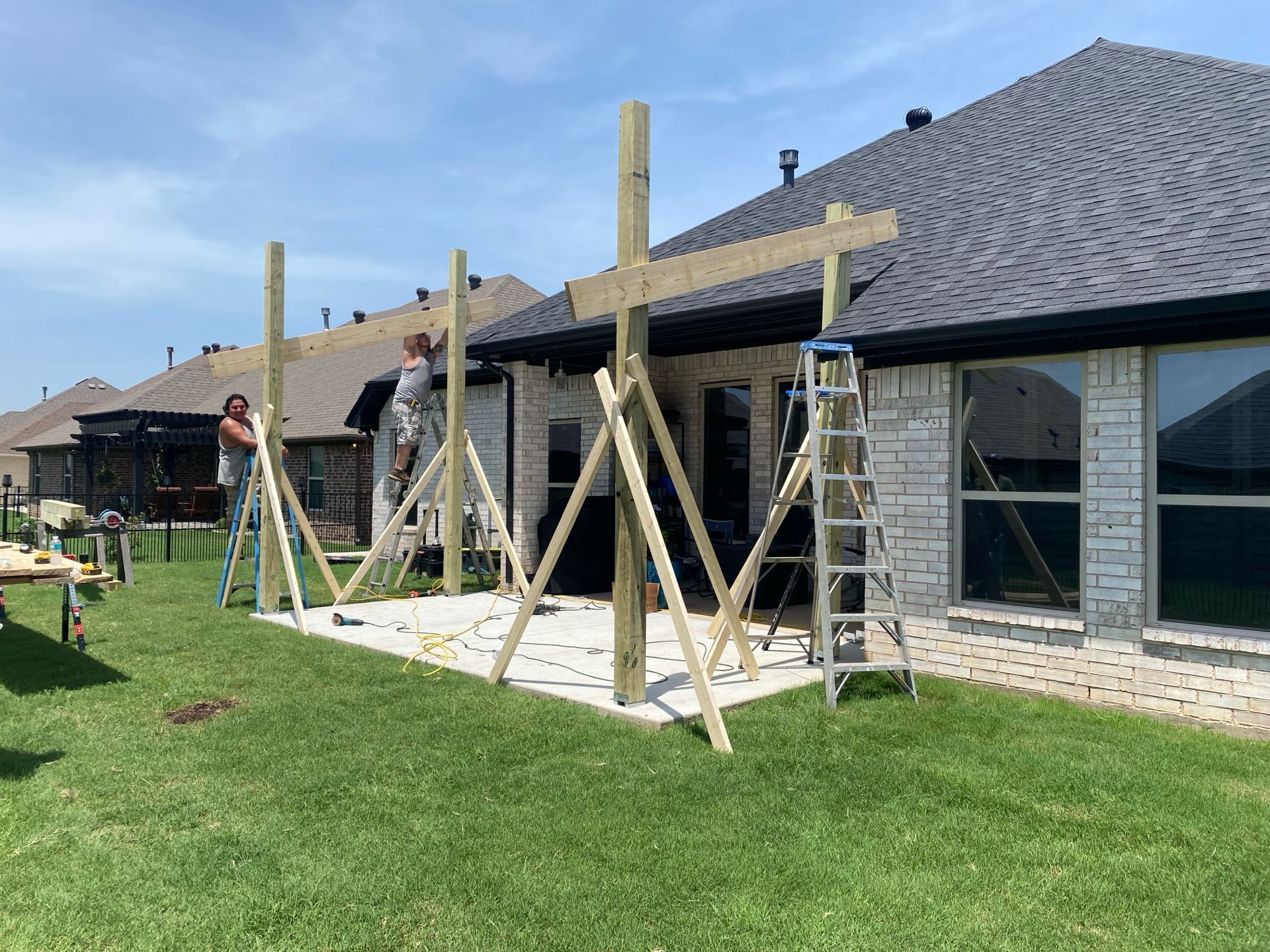 Two people building a wooden patio cover on a concrete slab next to a house.