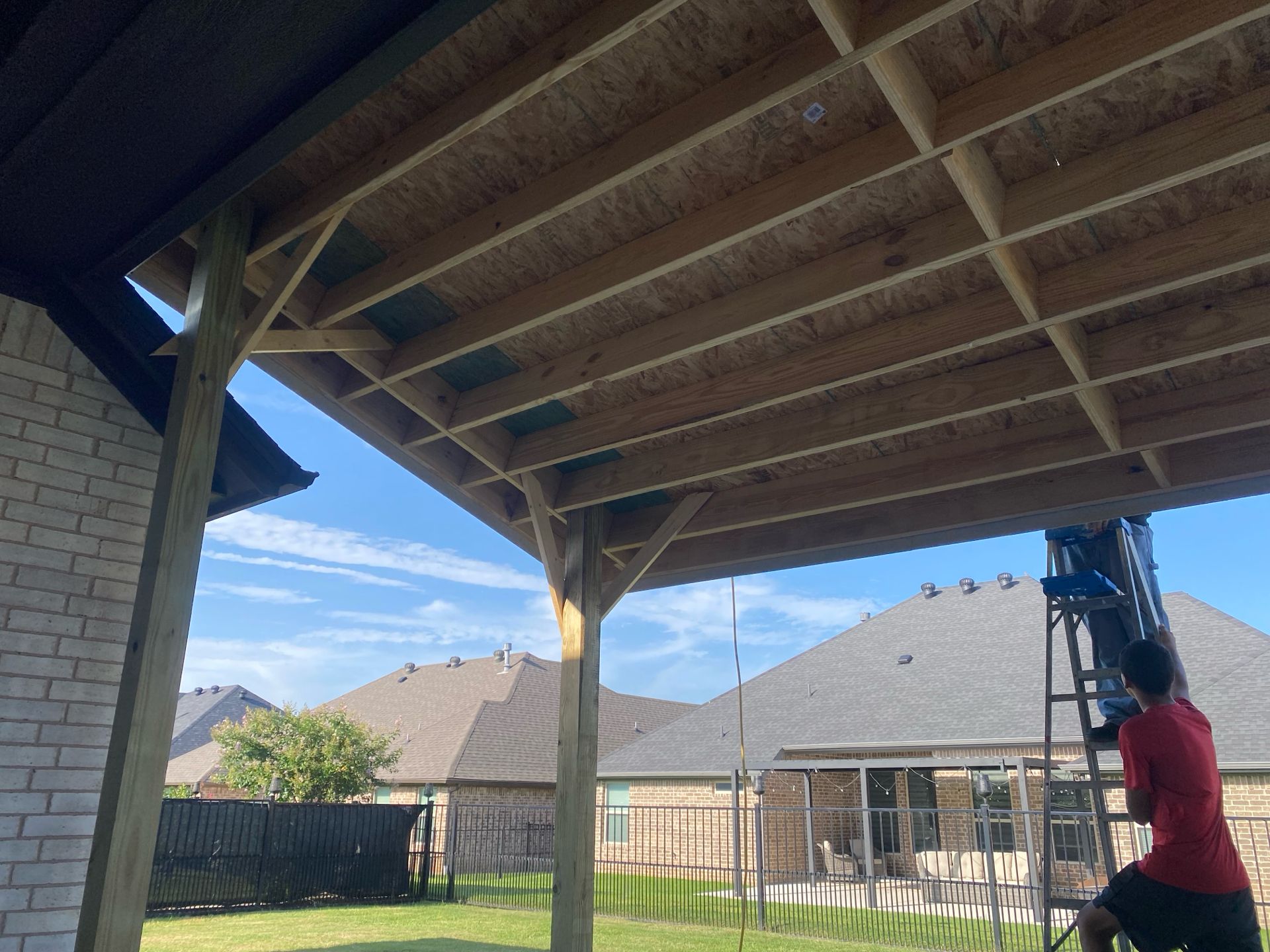 A man on a ladder works on a wooden patio roof extension. The sky is blue.