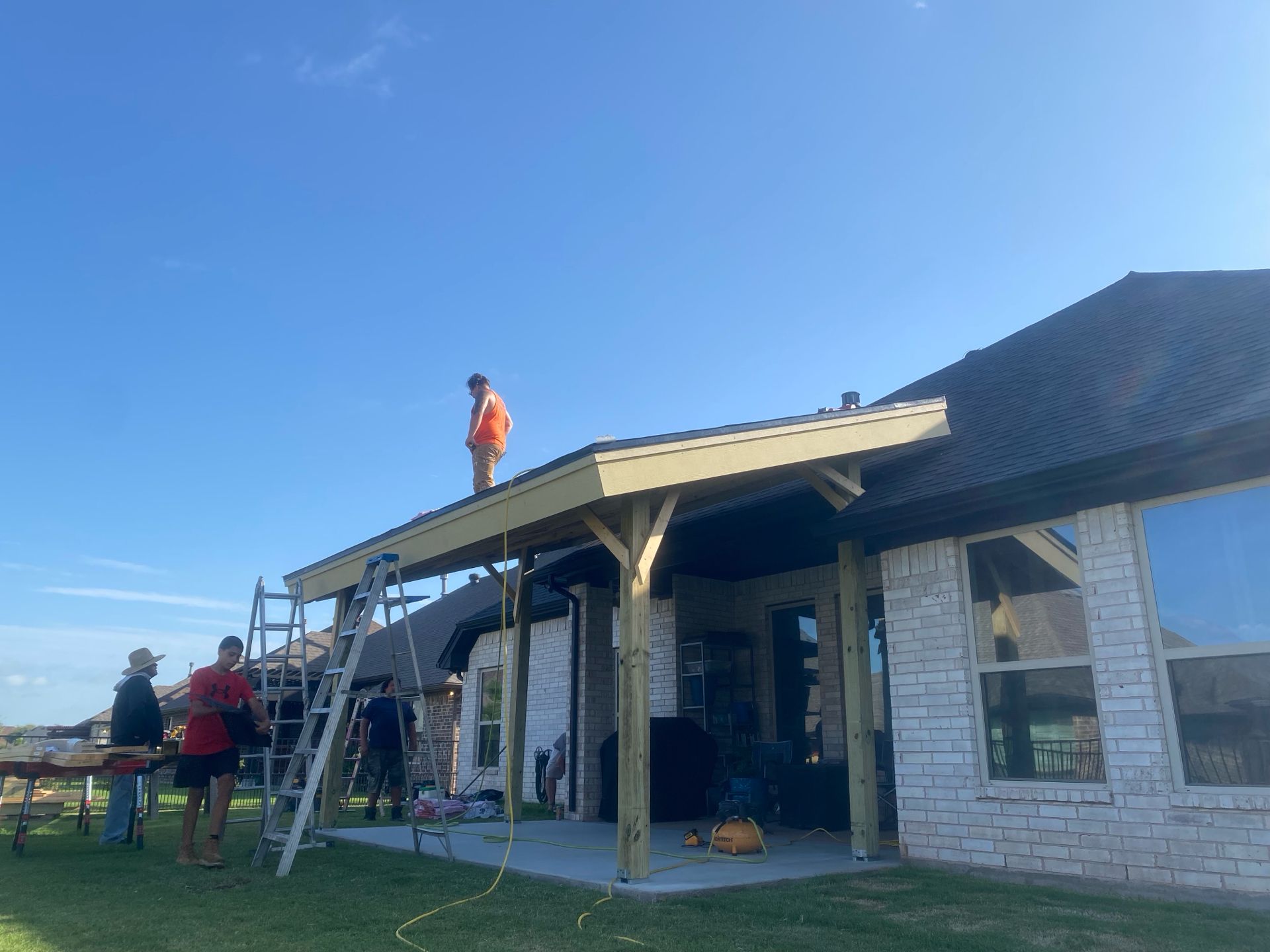 Construction workers building a covered patio extension on a house, under a clear blue sky.