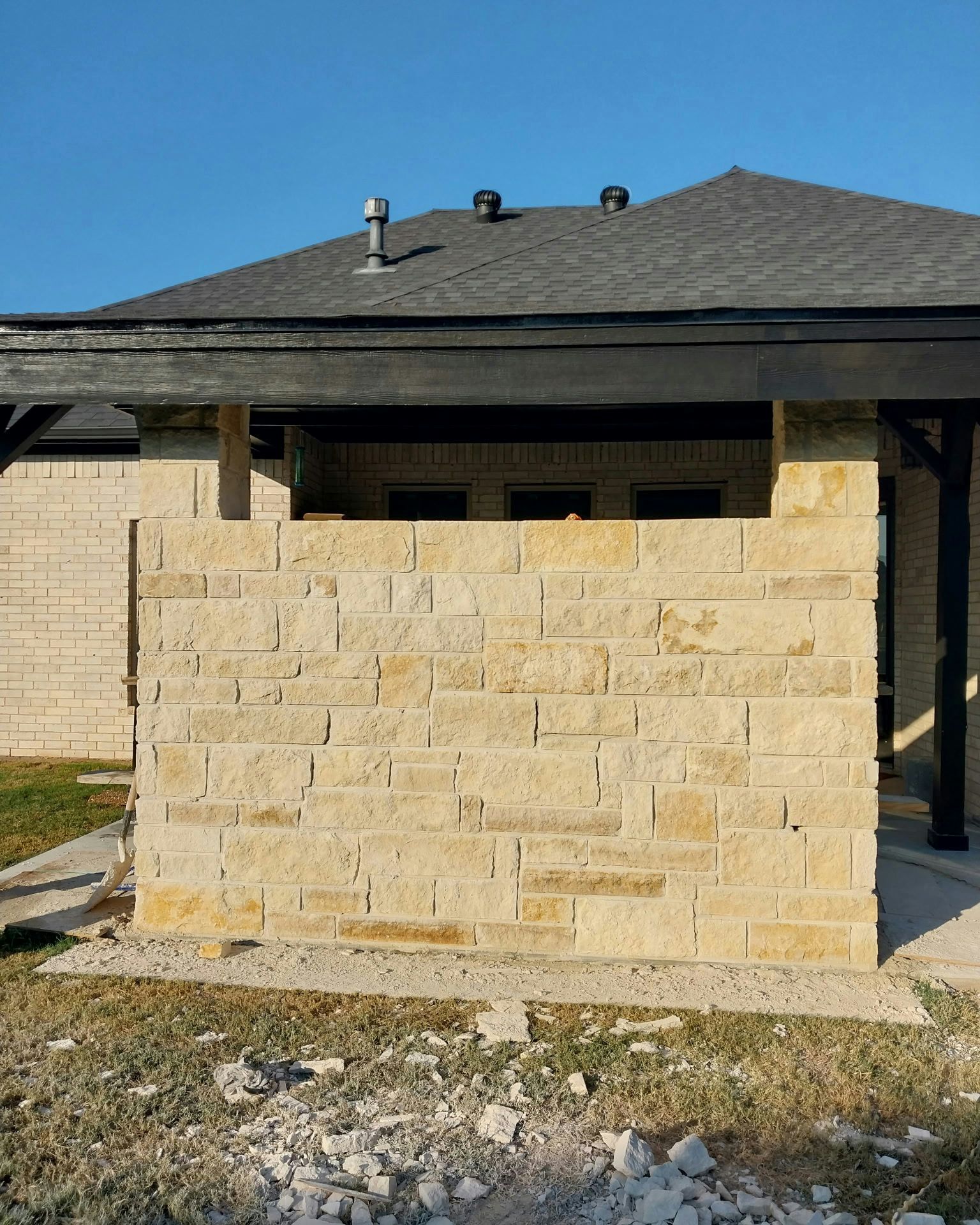 Stone wall under construction, tan bricks, dark roof, blue sky.