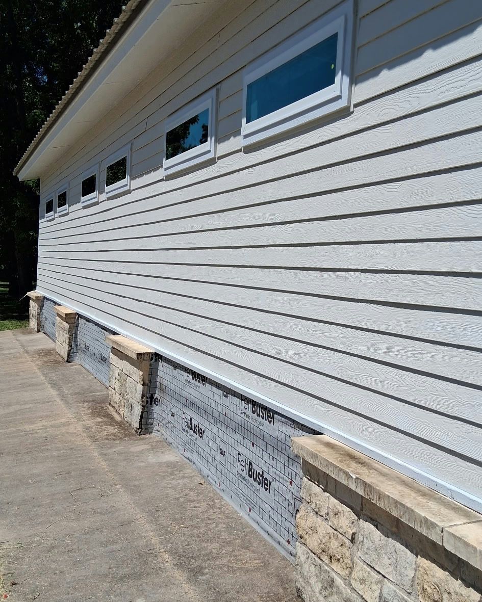 Exterior side of a building with light gray siding, small windows, and a stone and concrete foundation.