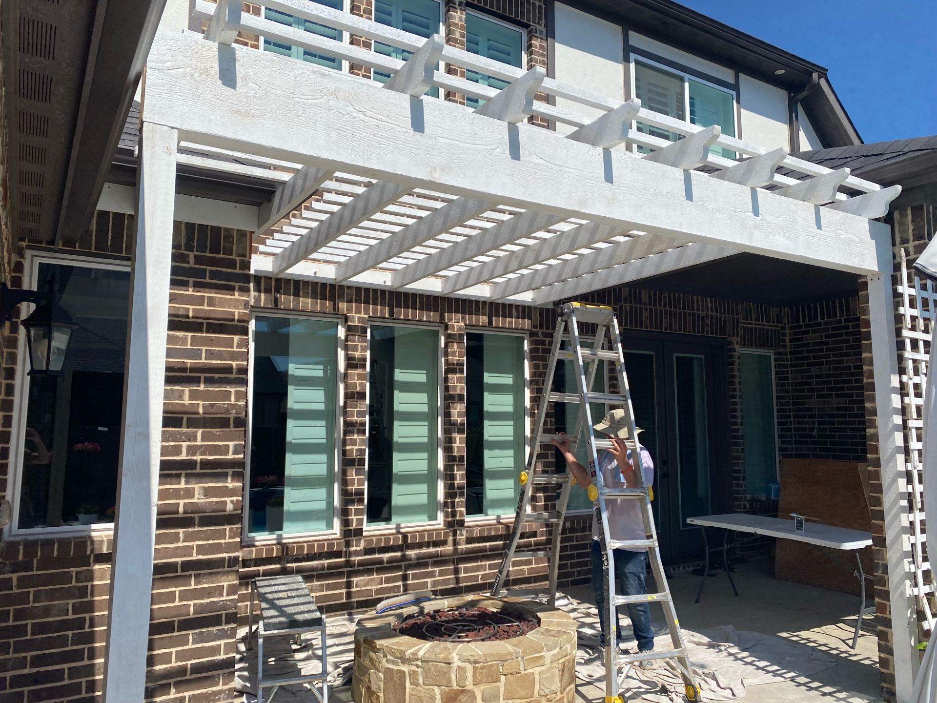 Person on ladder working on a white pergola attached to a brick house.