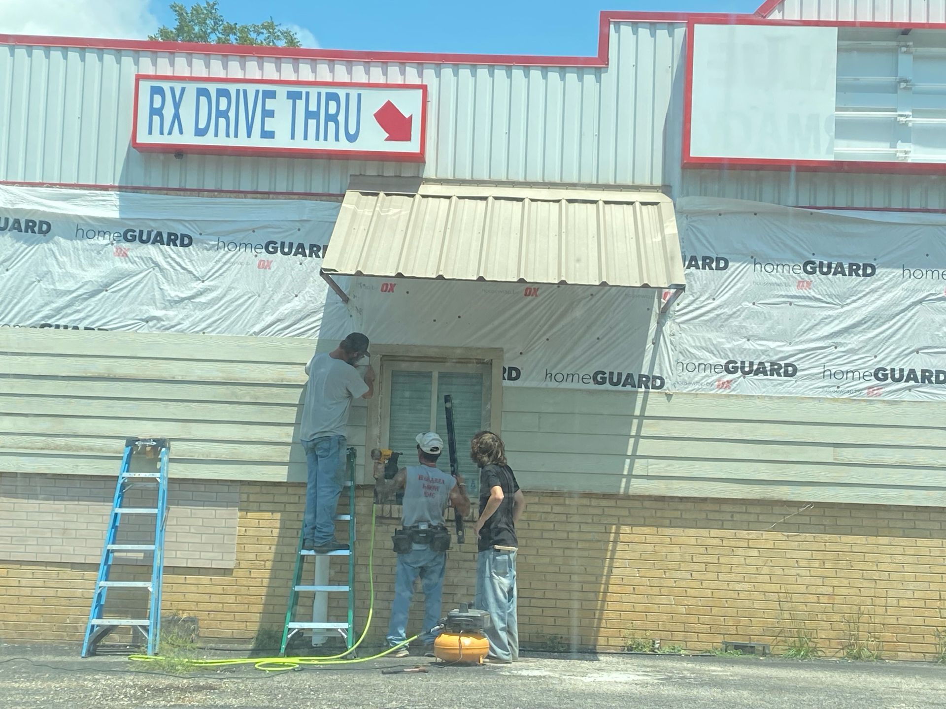 Construction workers installing siding on a building with a drive-thru pharmacy sign.