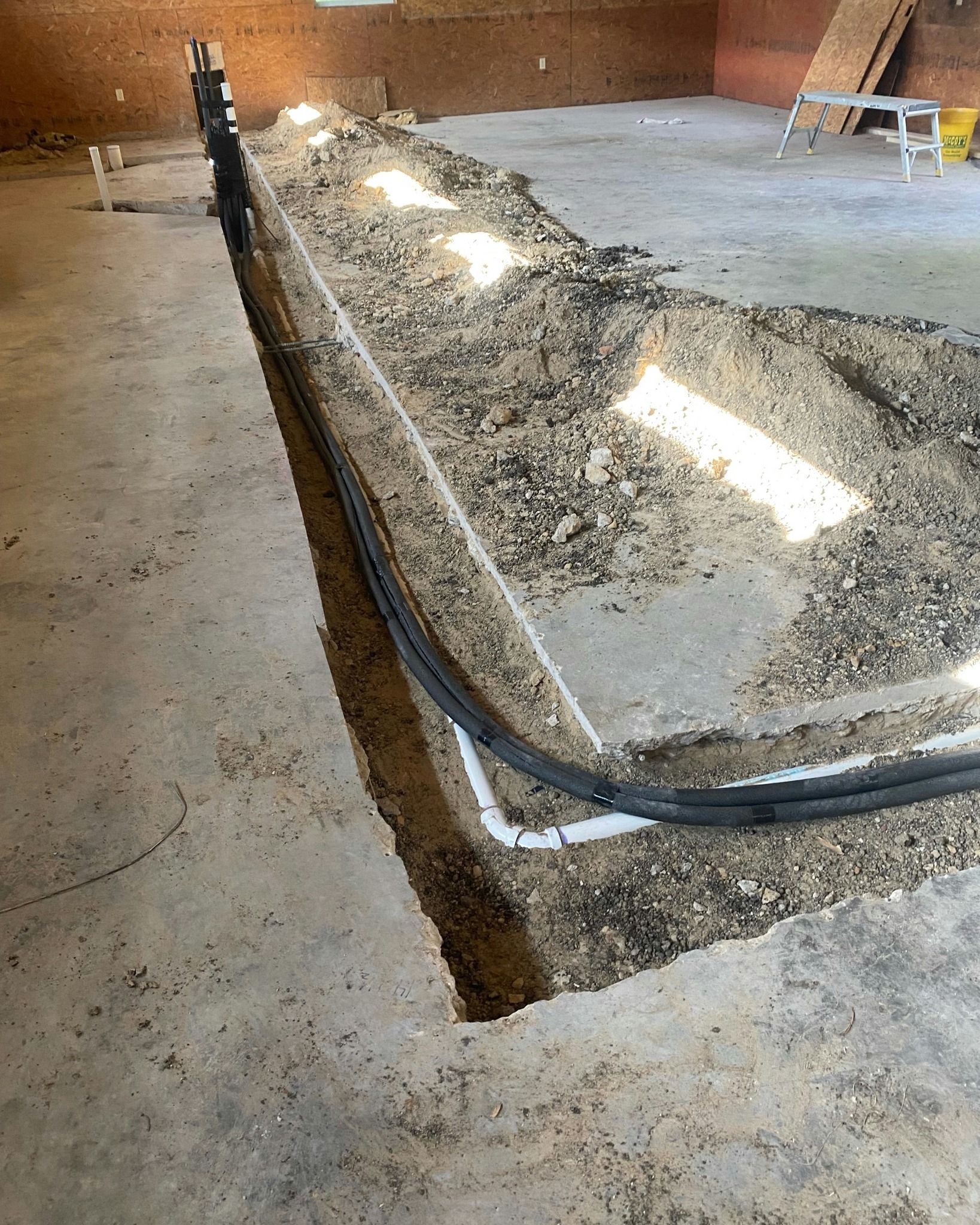 Trench dug in concrete floor, containing black conduit, filled with gravel. Indoor setting.