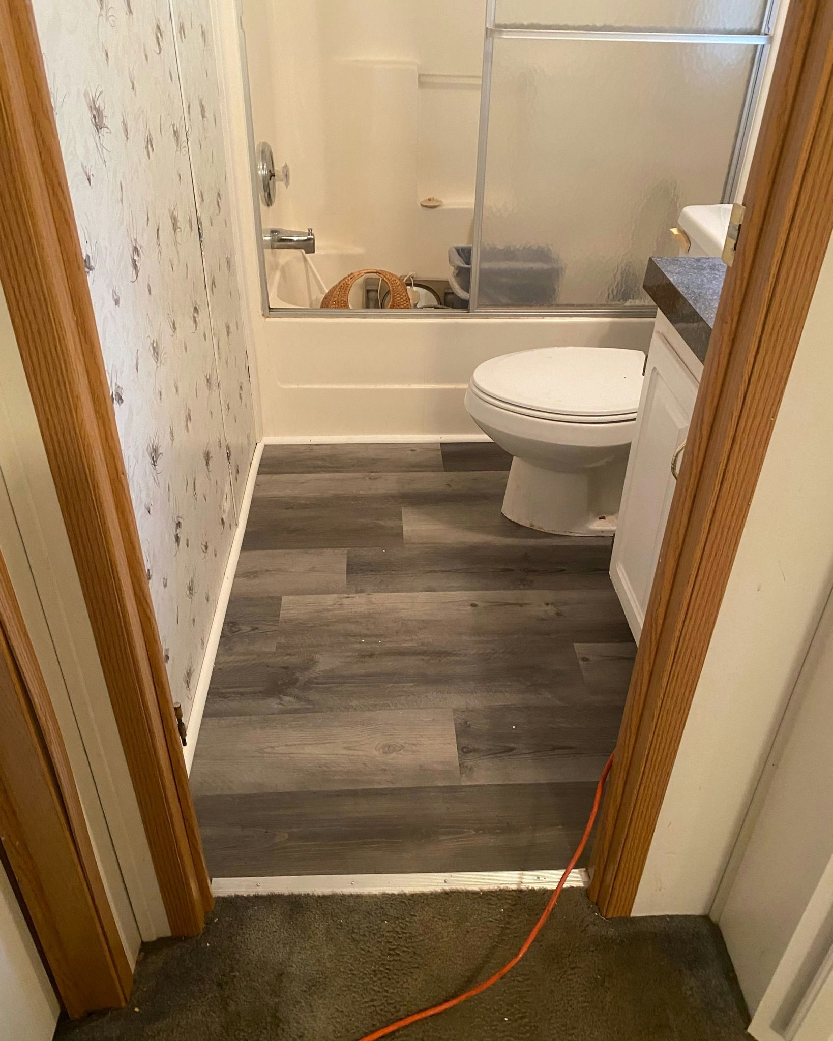 Bathroom with gray wood-look flooring, white toilet, shower/tub combo, and brown door frames.
