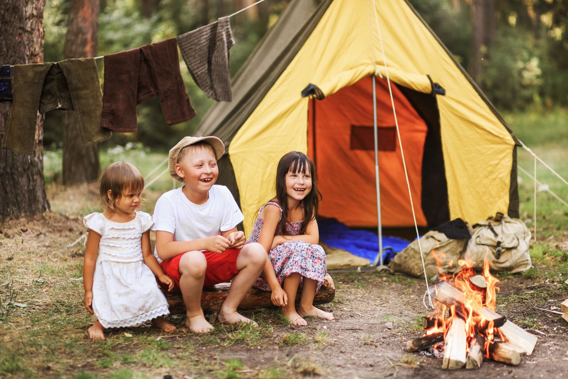 Three children sit by a yellow tent and campfire in a wooded campsite.