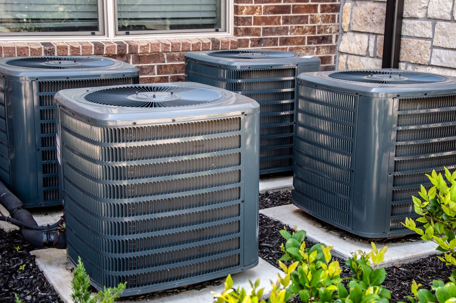 Four gray air conditioning units outside, near brick and stone walls, with green bushes.