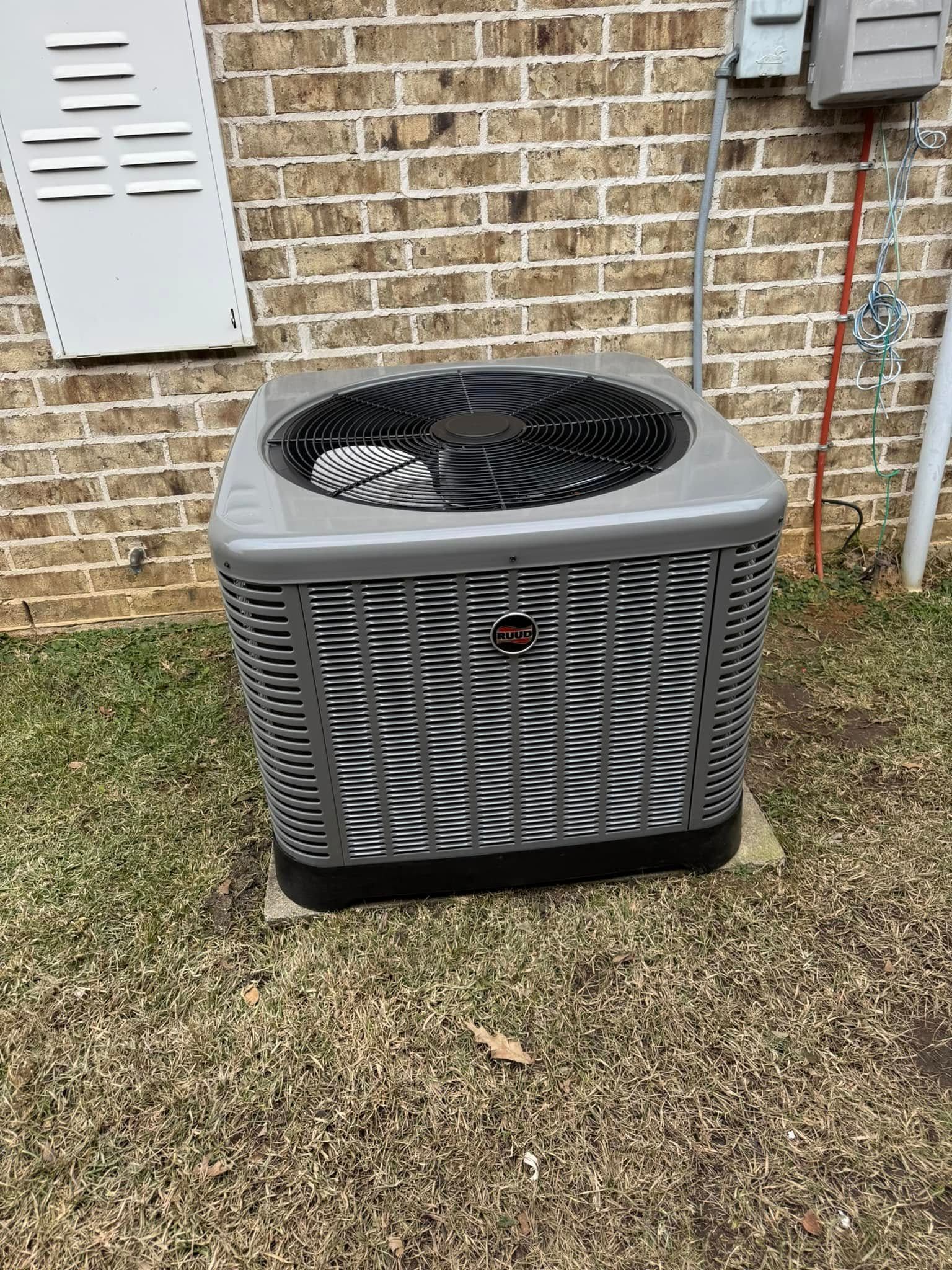 An outdoor air conditioning unit sits on a concrete pad near a brick wall and a utility box.