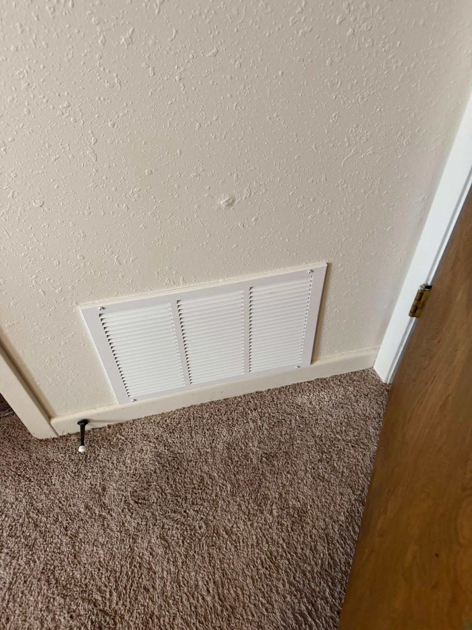 White air vent on textured wall above brown carpet next to a door frame.