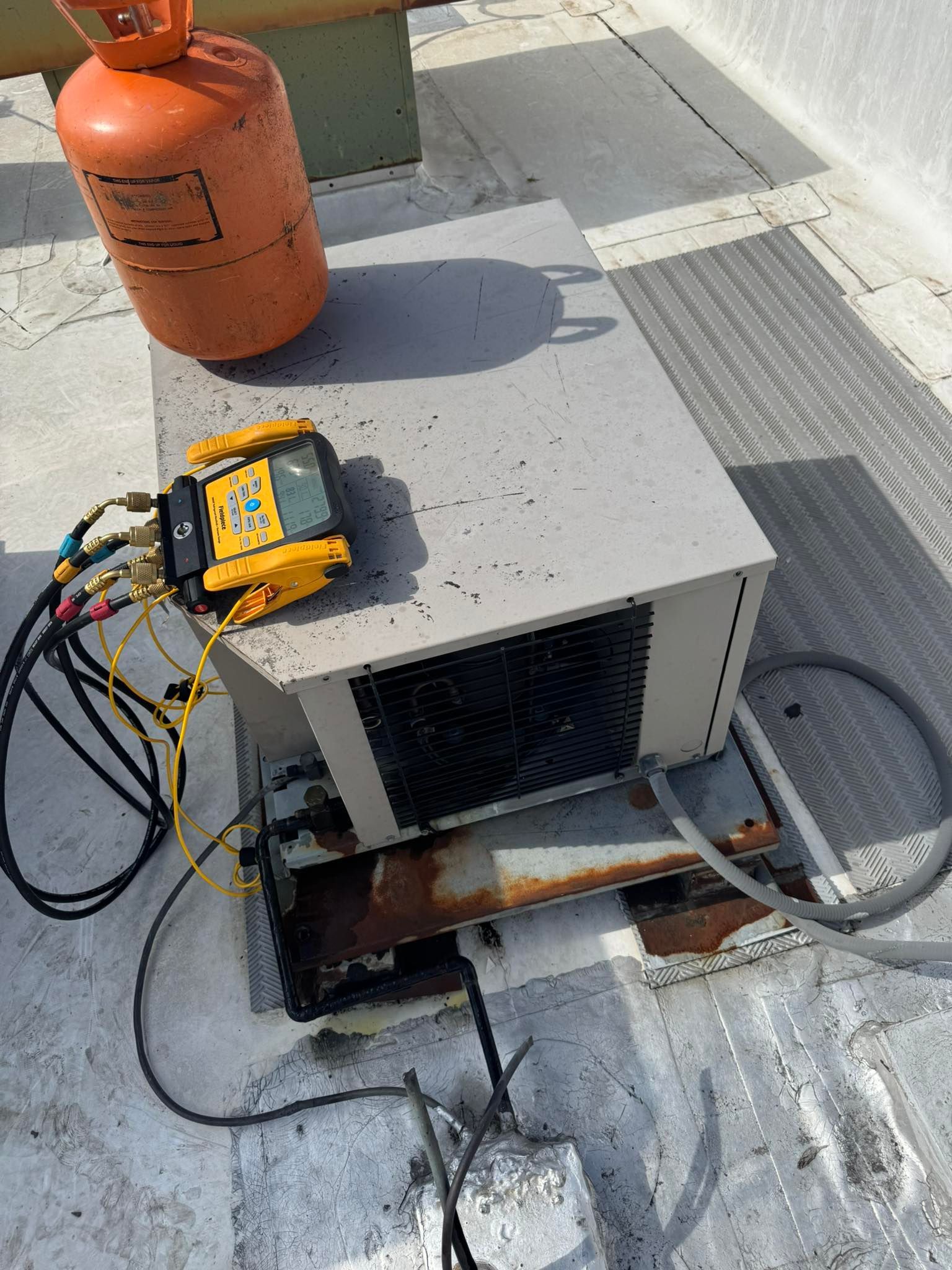 HVAC equipment on a rooftop; gauge manifold connected, orange refrigerant tank nearby.