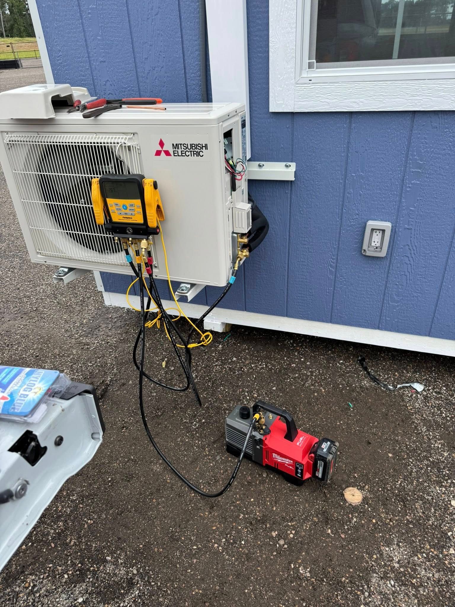 HVAC technician servicing a Mitsubishi mini-split unit on a blue building; vacuum pump in use.