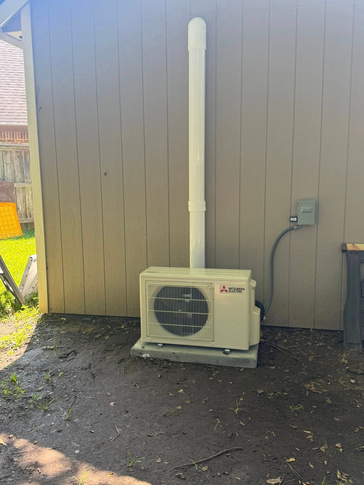 An air conditioning unit is installed on a concrete pad against a beige building, with a white exhaust pipe.