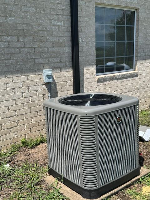 An air conditioning unit sits next to a light brick wall, with a window and a black pipe above it.