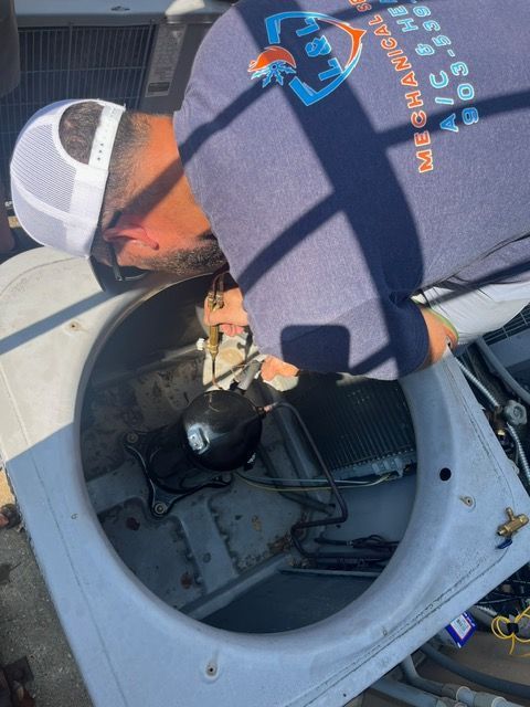 HVAC technician repairs an air conditioner unit. He wears a hat and sweatshirt. The unit is metal and gray.