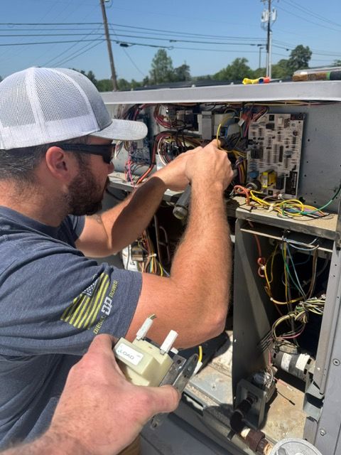 Man in hat repairs HVAC unit, focused, outdoors, bright day. Another hand holds a part.