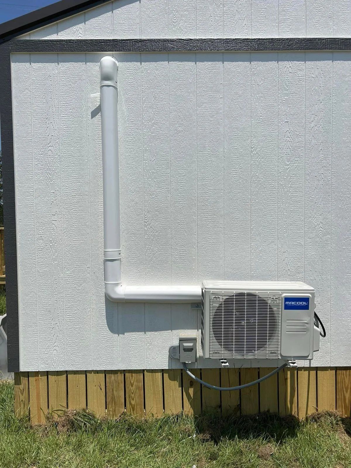 White air conditioning unit mounted on a textured white building exterior with drainage pipe, next to wooden base.