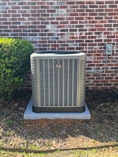 A tan outdoor air conditioning unit sits on a concrete pad against a brick wall next to a green shrub.
