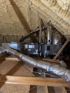HVAC unit and silver insulated ductwork installed in an attic with spray foam insulation on the rafters.