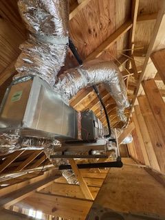 A silver rectangular metal duct with attached flexible insulated tubing mounted to wooden attic rafters.