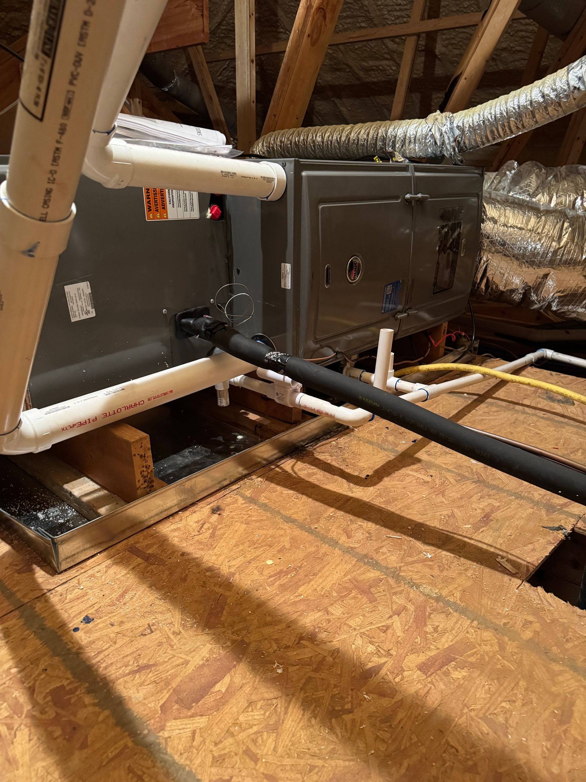 HVAC unit in an attic with white pipes and black hose resting on wood beams.