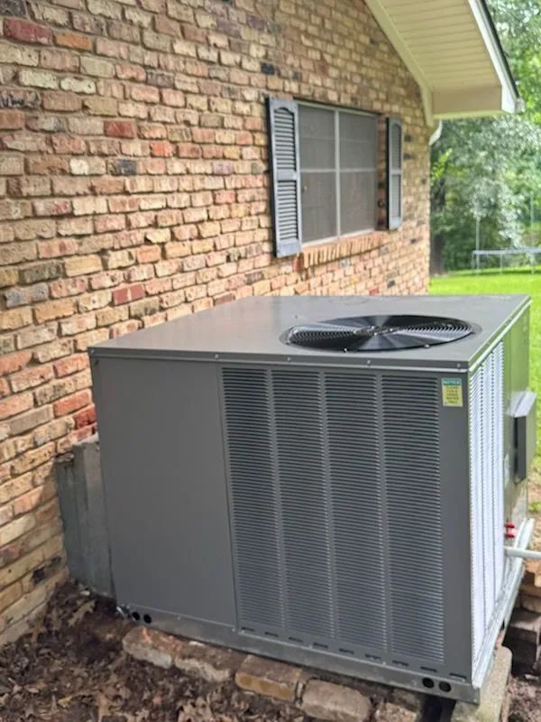 Gray air conditioning unit next to a brick wall under a house eave.