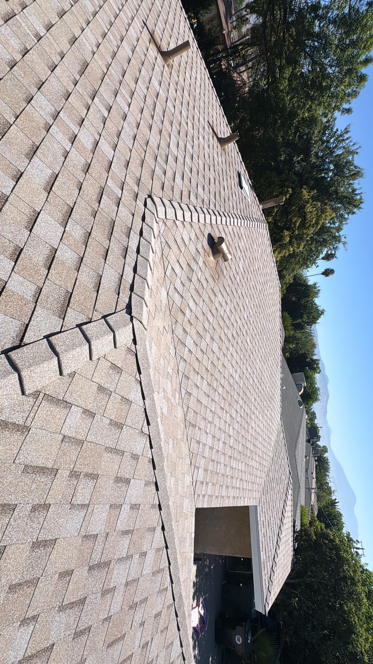 Overhead view of a roof with gray shingles, a small chimney, and a blue sky.