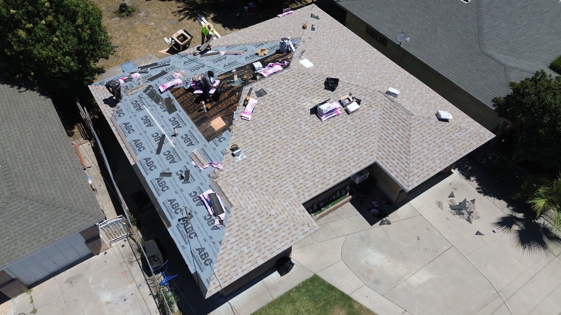 Aerial view of a house undergoing a roof replacement; workers are present, laying down shingles and protective layers.