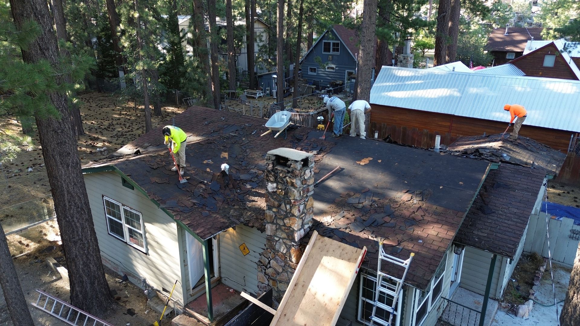 Workers replace the roof of a cabin in a wooded area, with a chimney and surrounding trees.