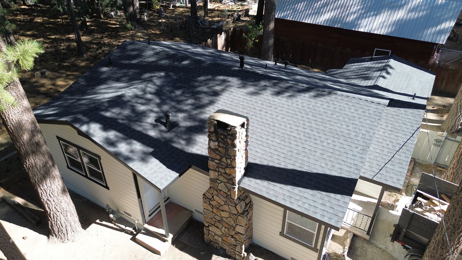 Cabin with gray shingle roof and stone chimney, surrounded by trees.