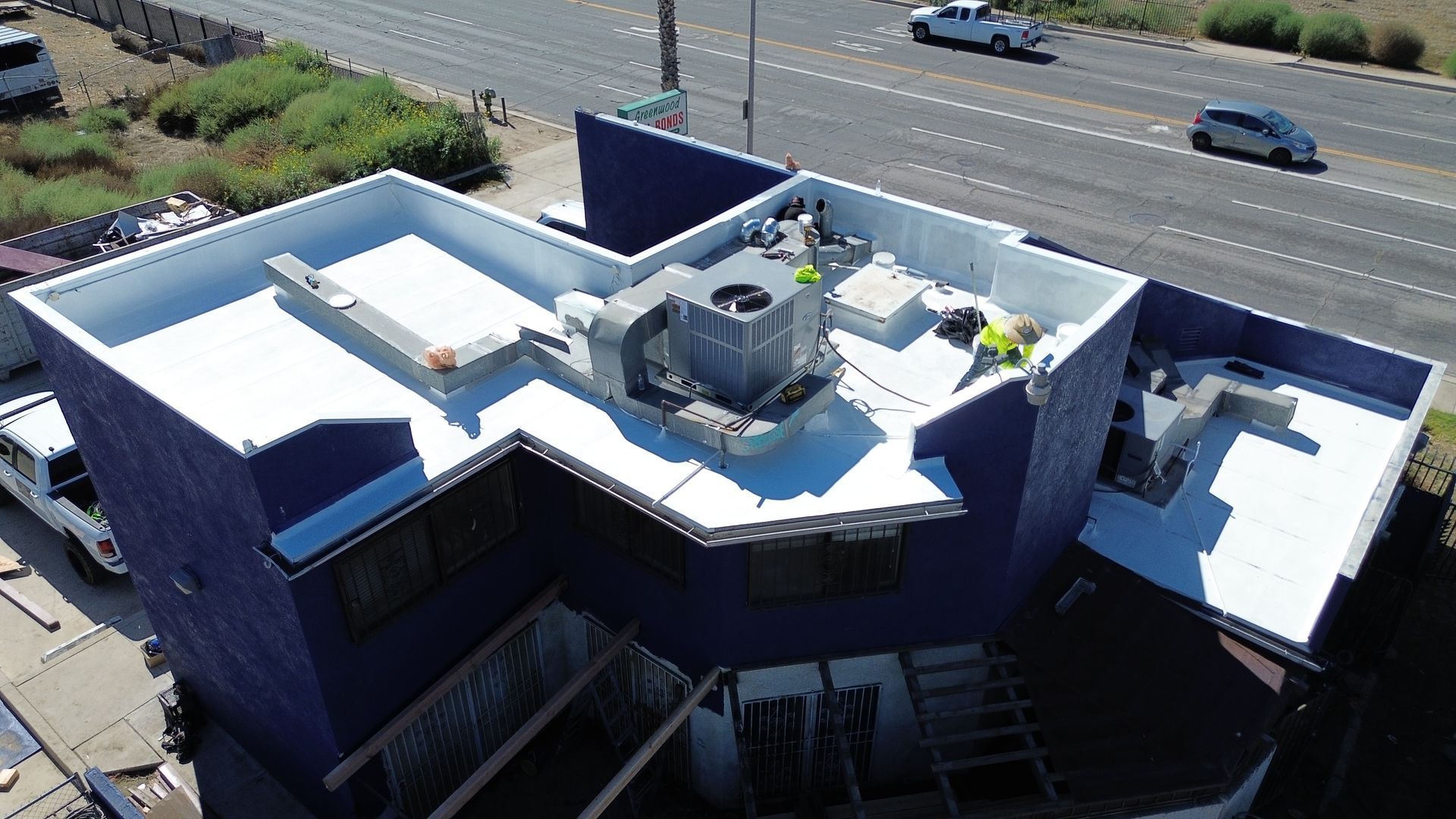 A rooftop view of a blue building with white roofing. Workers are on the roof near HVAC units. Cars are driving nearby.