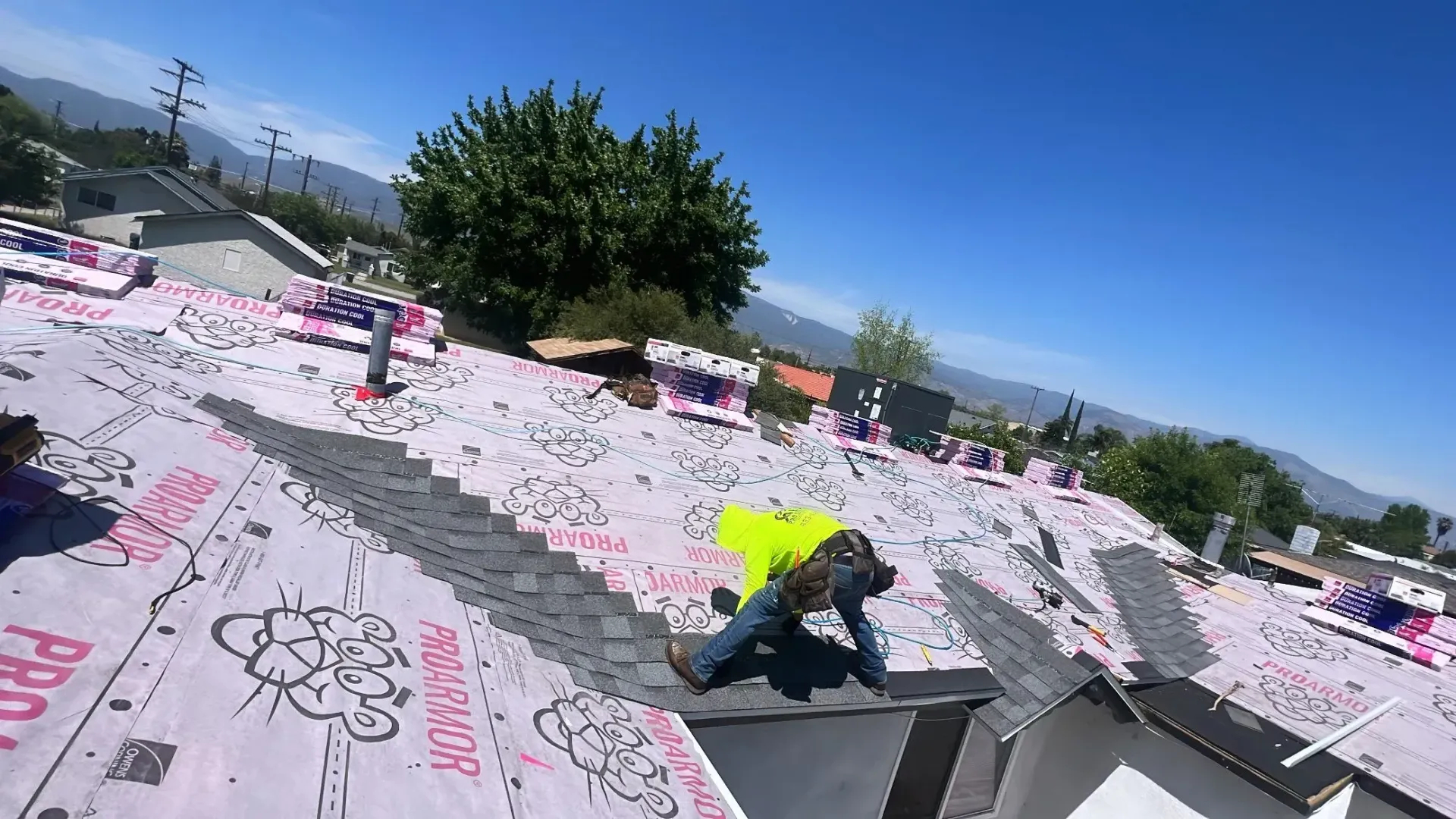 Roofer on a roof working under a bright blue sky. They are wearing a neon yellow vest.