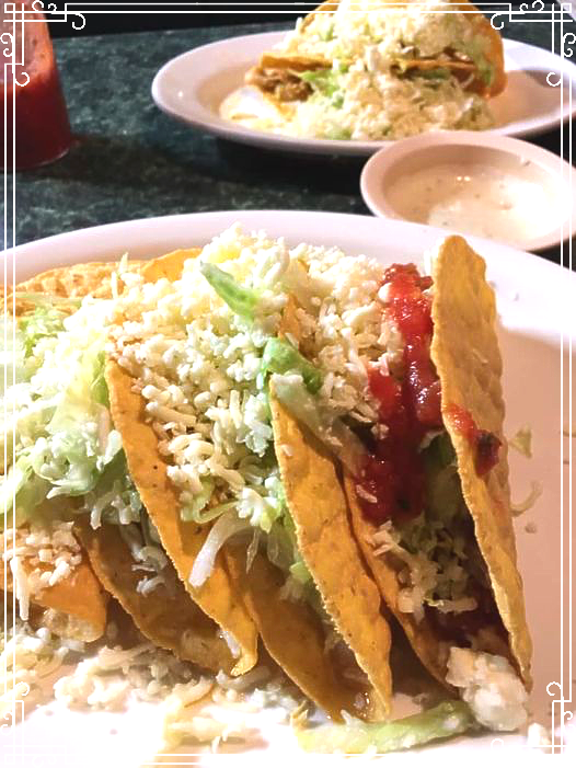 Hard shell tacos with lettuce, cheese, and salsa; another dish in the background.