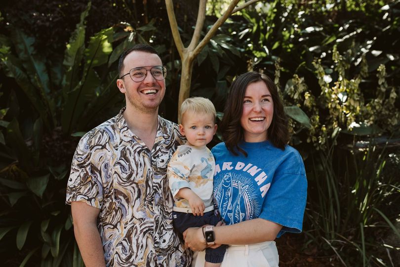 A smiling couple stands outdoors with a small child in their arms.