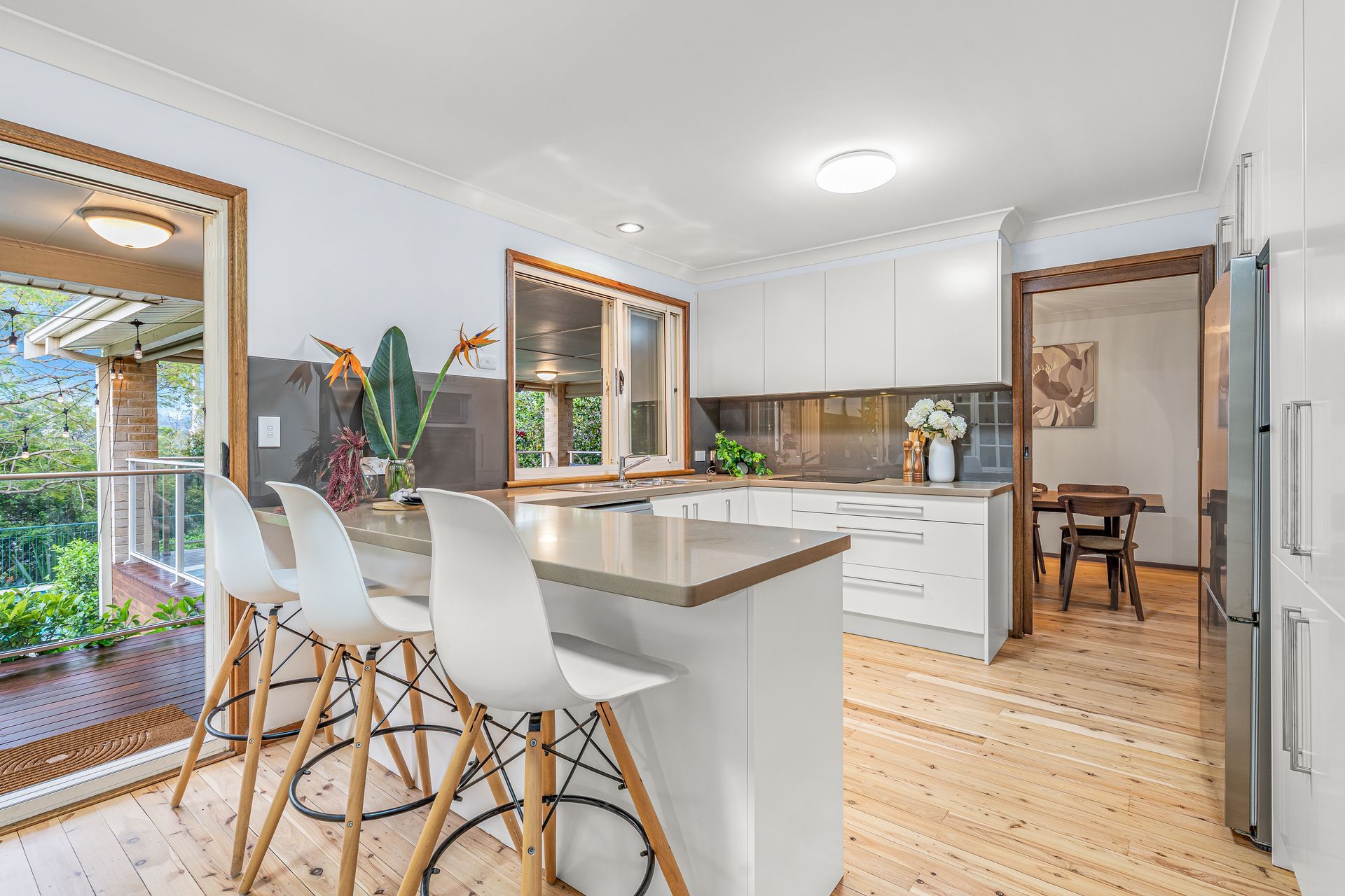 A bright kitchen with white cabinets, a light wood floor, a stone island with three white stools, and an outdoor balcony — Brightside Buyers Agency in Dundas, NSW