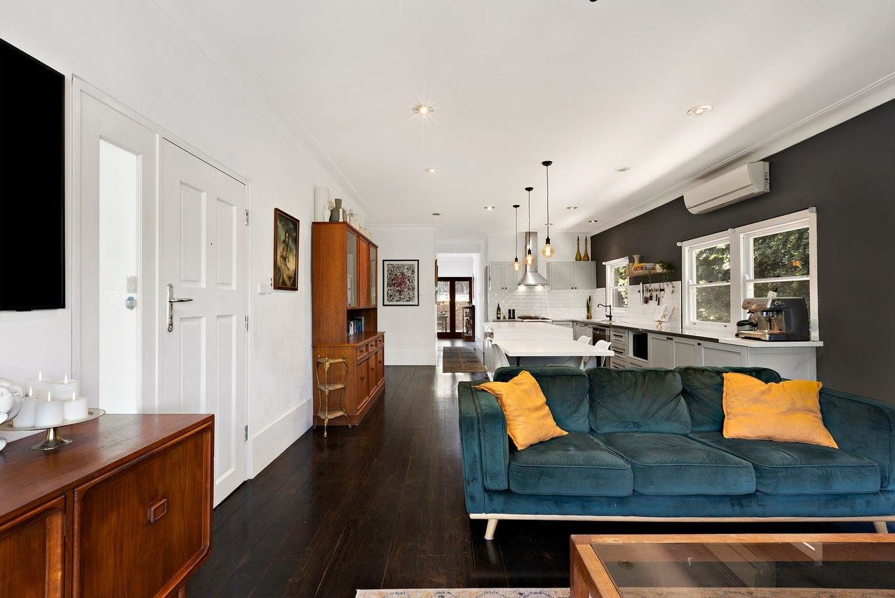 A modern open-plan living room with a dark blue sofa, yellow cushions, dark wood floors, and a kitchen in the background — Brightside Buyers Agency in Dundas, NSW