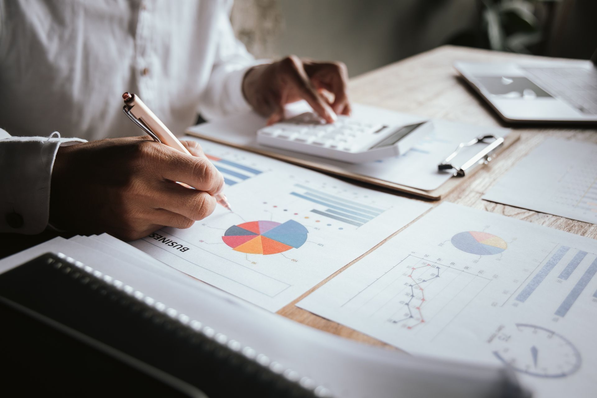 Person using a calculator, writing on charts with colorful pie graph and data, on a desk.