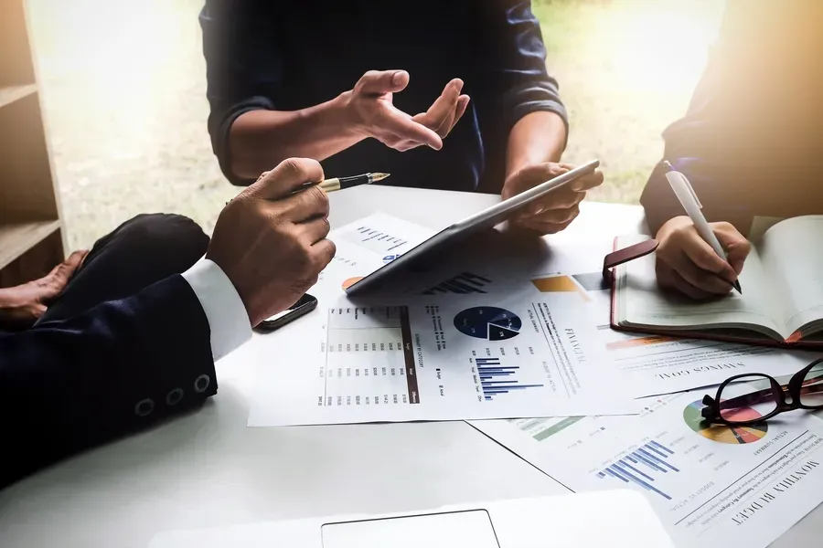 Three people at a table reviewing financial data, including charts, tablet, and notebook.