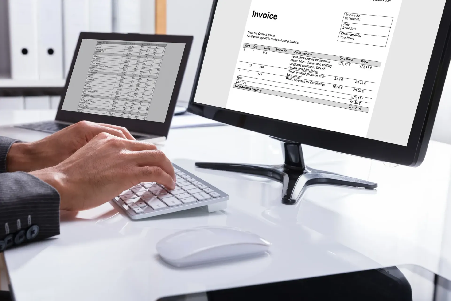 Person typing on a keyboard, with two computer screens showing invoices and data on a white desk.