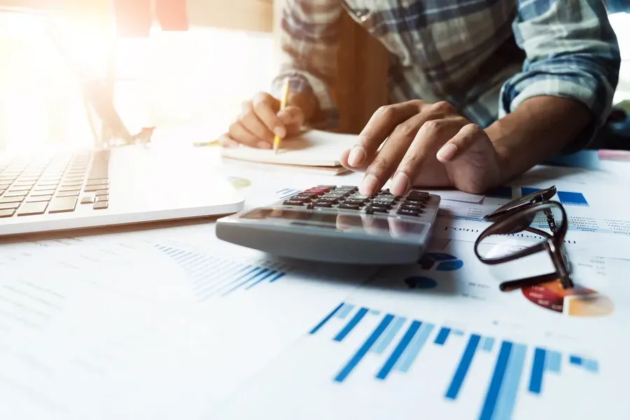 Person using a calculator, notebook, and laptop, with charts and glasses on the desk.
