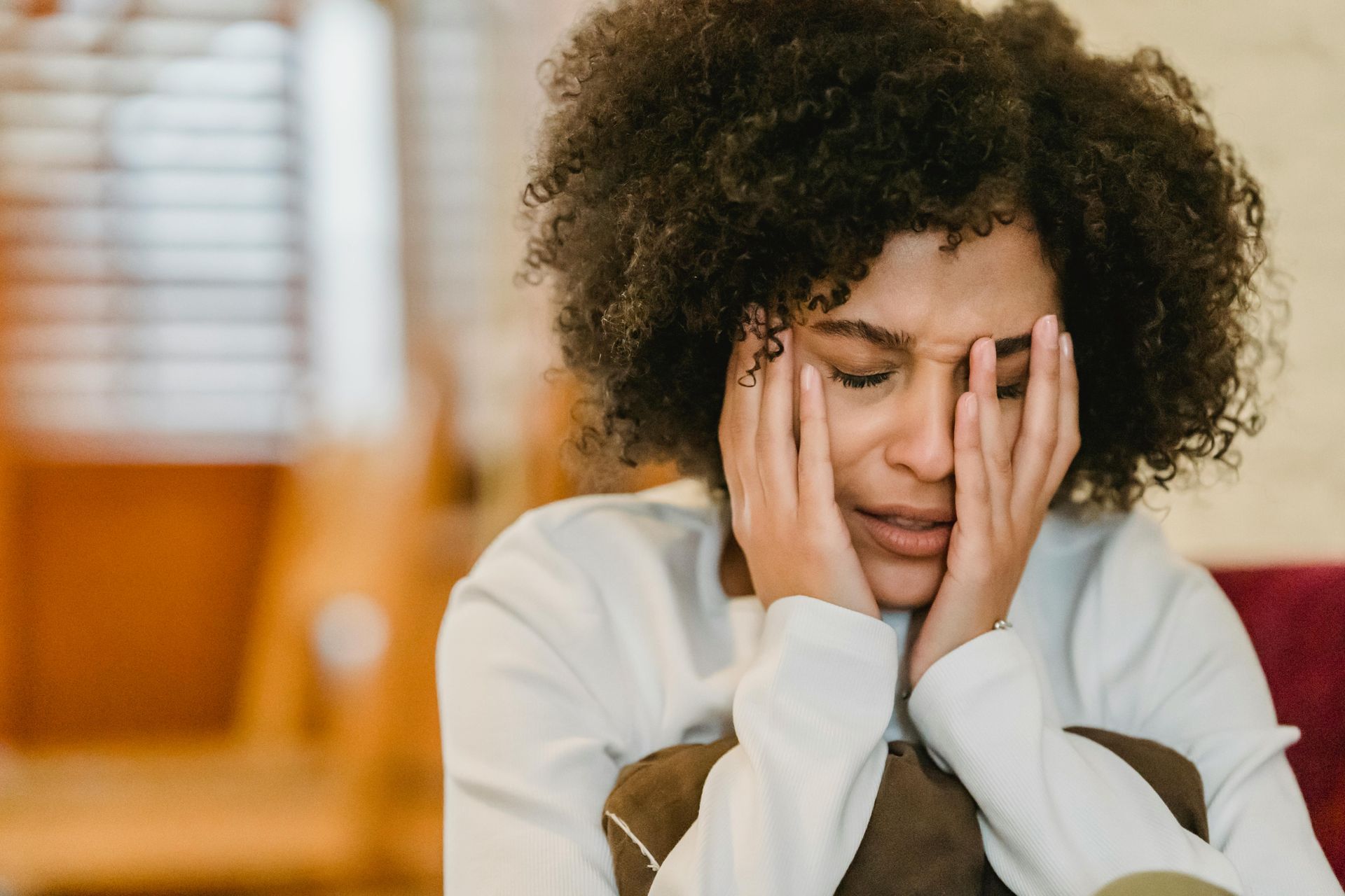 A woman with curly hair is sitting on a couch covering her face with her hands.