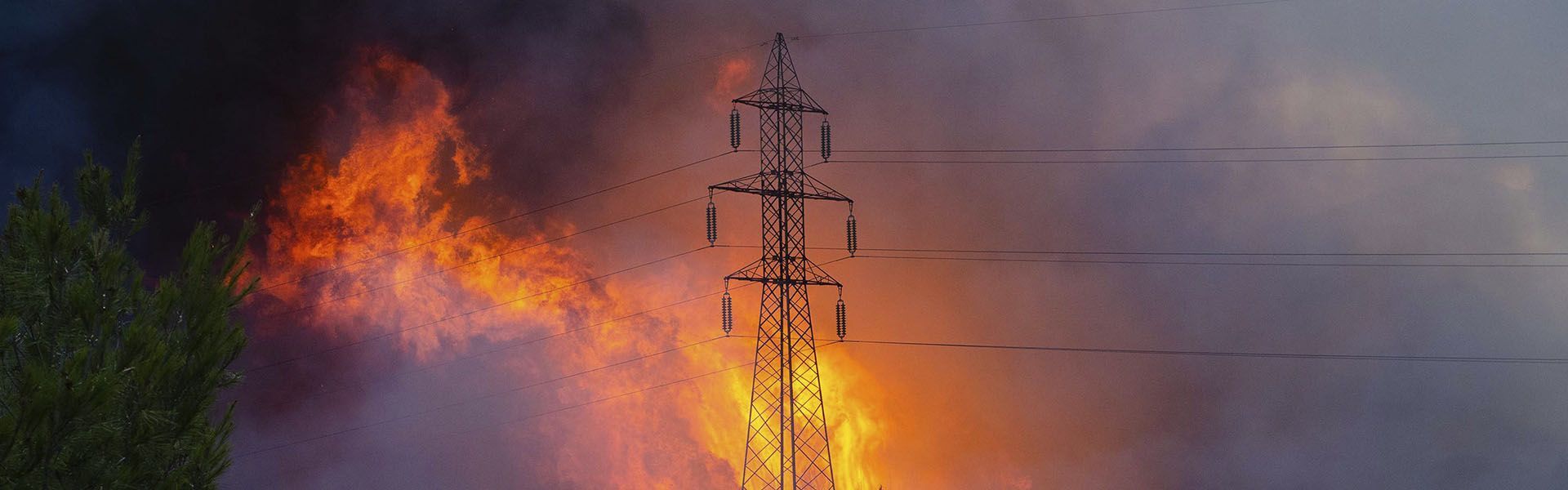 Foto contendo uma torre de transmissão de energia elétrica envolta por chamas e fumaça escura.