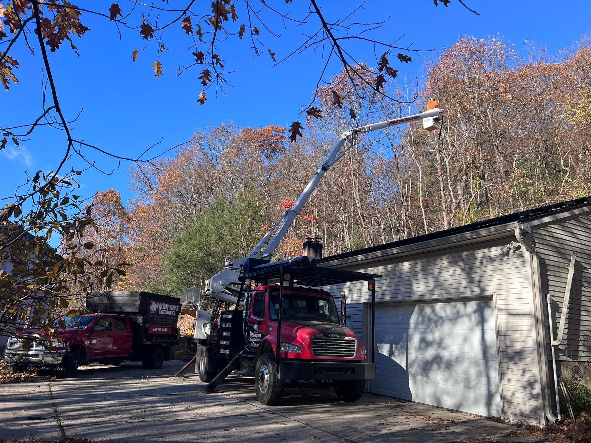 Utility truck with extended boom parked beside a garage, with another truck in the driveway under autumn trees.