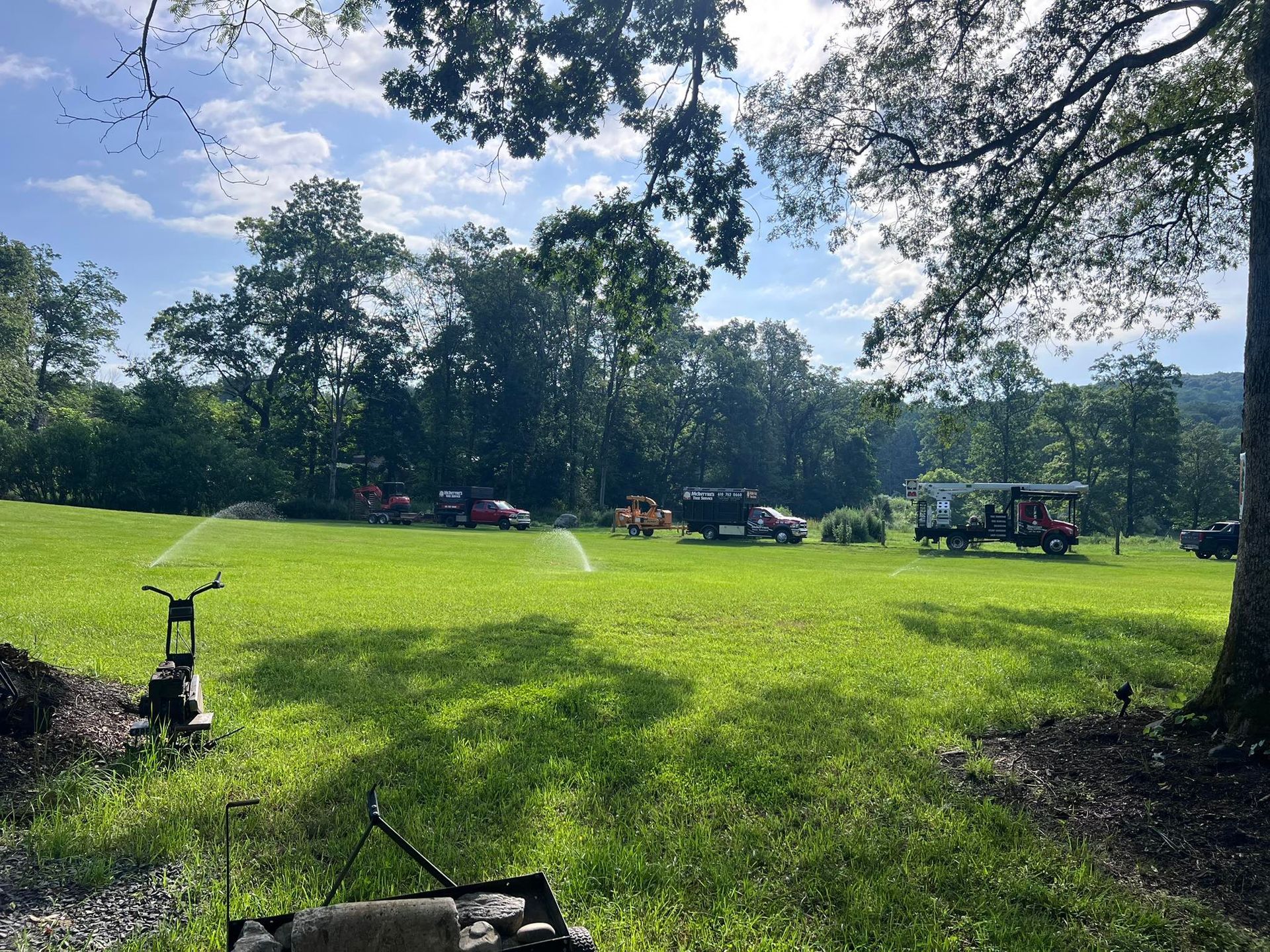Green field with sprinklers watering the grass, vehicles parked in the distance, trees and blue sky above.