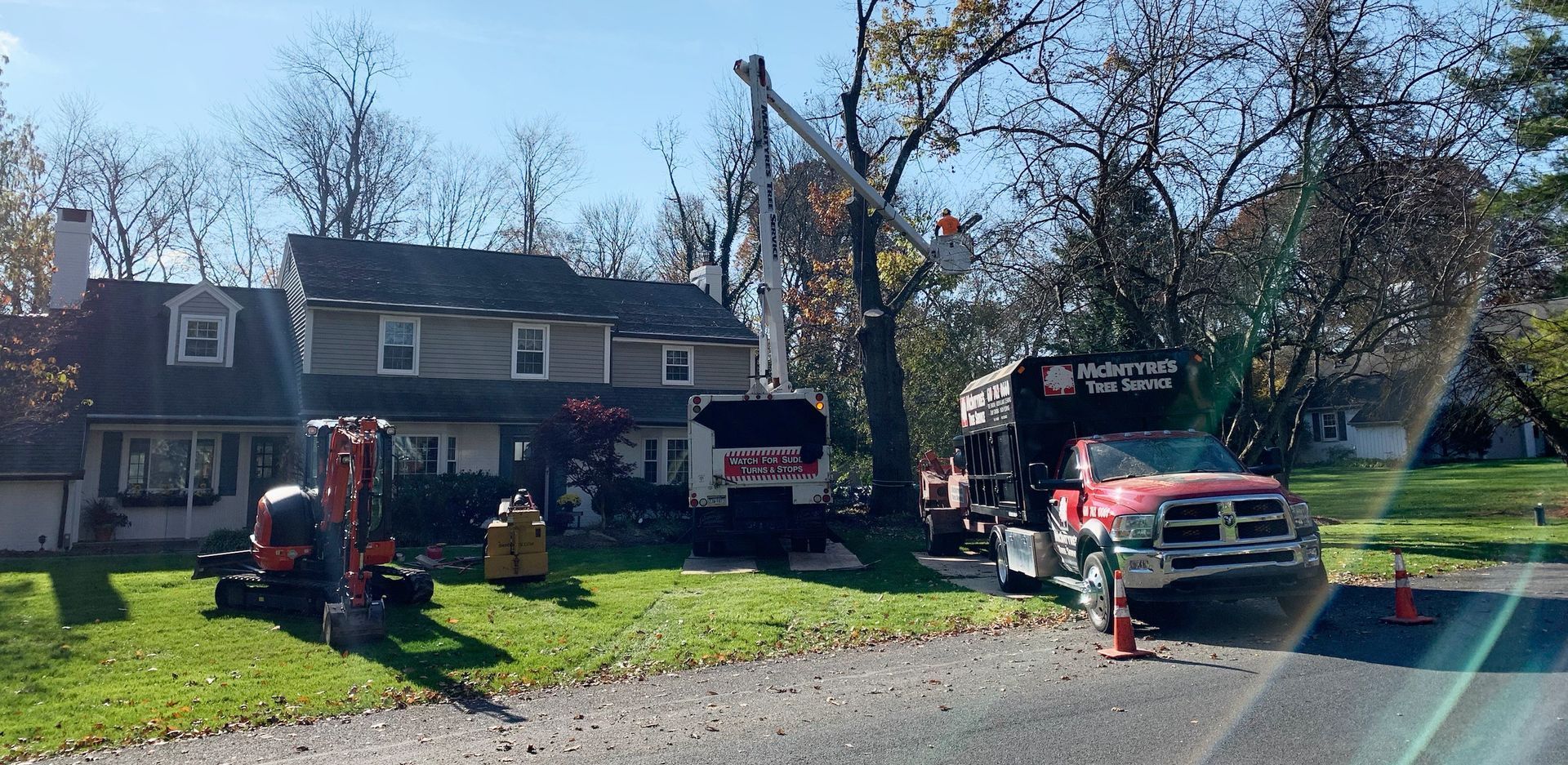 Tree service in progress: a bucket truck trimming a tree next to a house; a truck parked on the street.