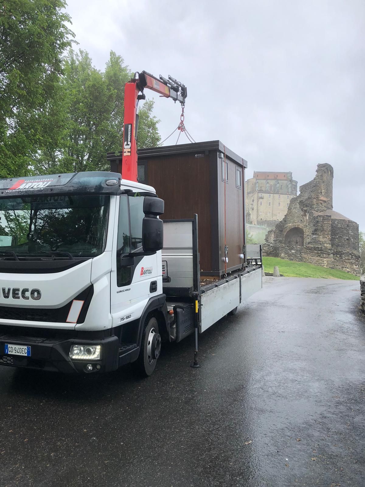 Un camion bianco a pianale con gru rossa trasporta una grande cassa marrone su una strada bagnata vicino a rovine in pietra.
