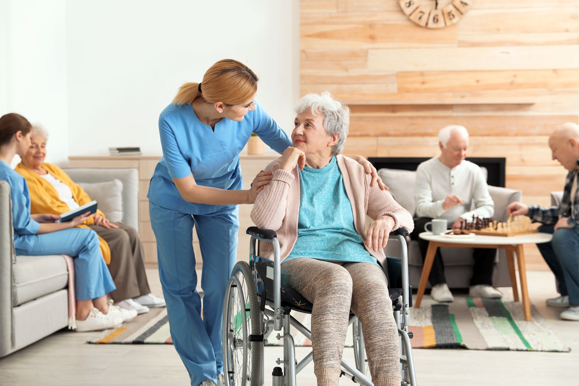 Nurse assisting a woman in a wheelchair; others sit and play games in a senior living facility.