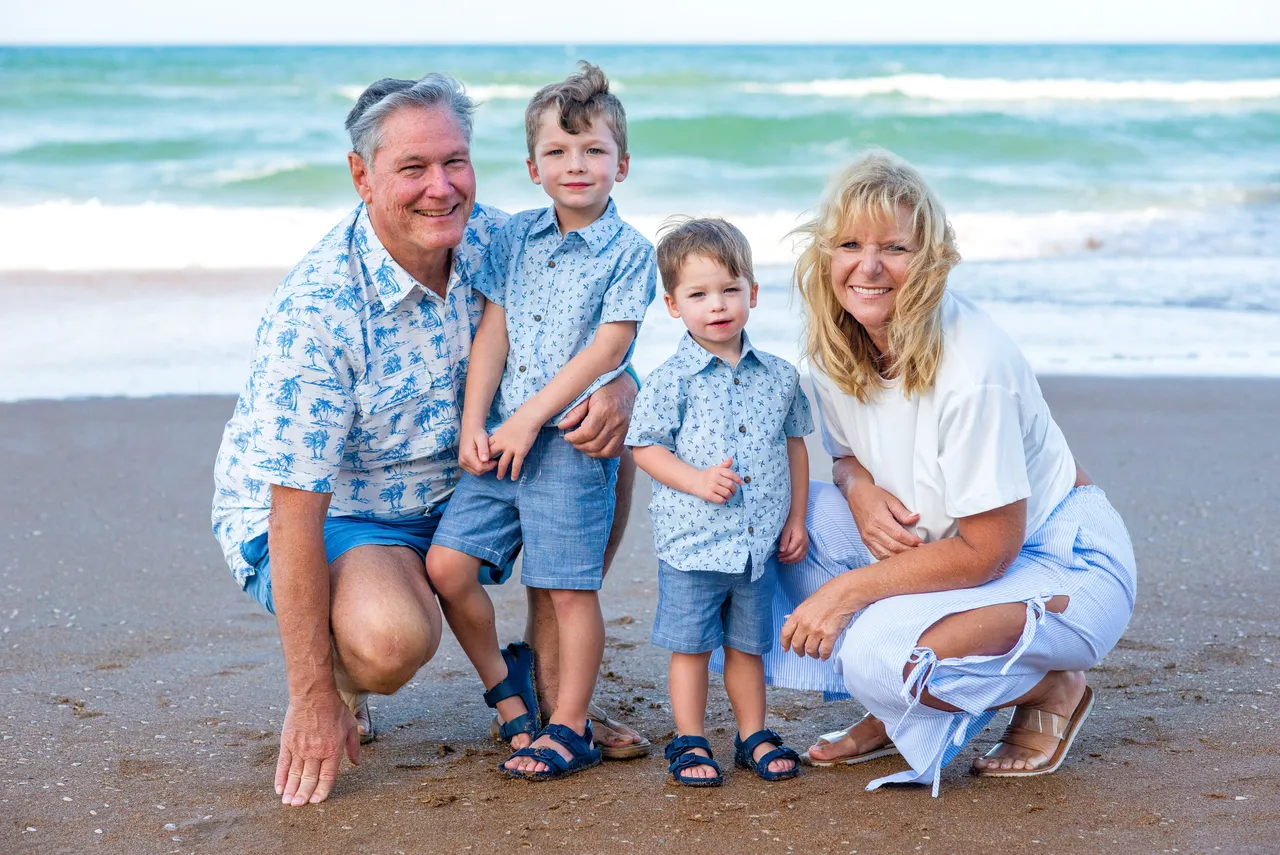 A family of four poses on a sandy beach by the ocean, all smiling and dressed in blue and white coordinating outfits.