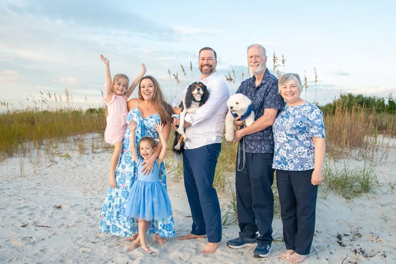 A multi-generational family and two dogs pose together on a sandy beach at sunset.
