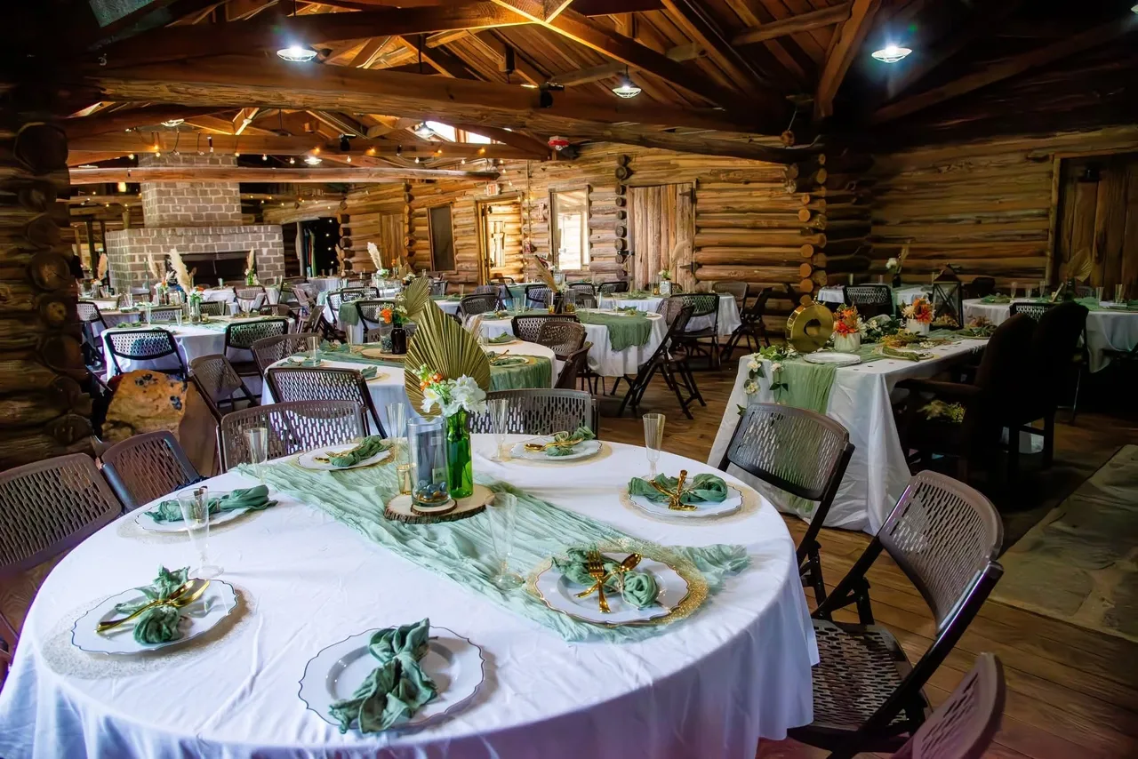 A rustic log cabin event hall set for a wedding or dinner with white round tables, green runners, and floral centerpieces.