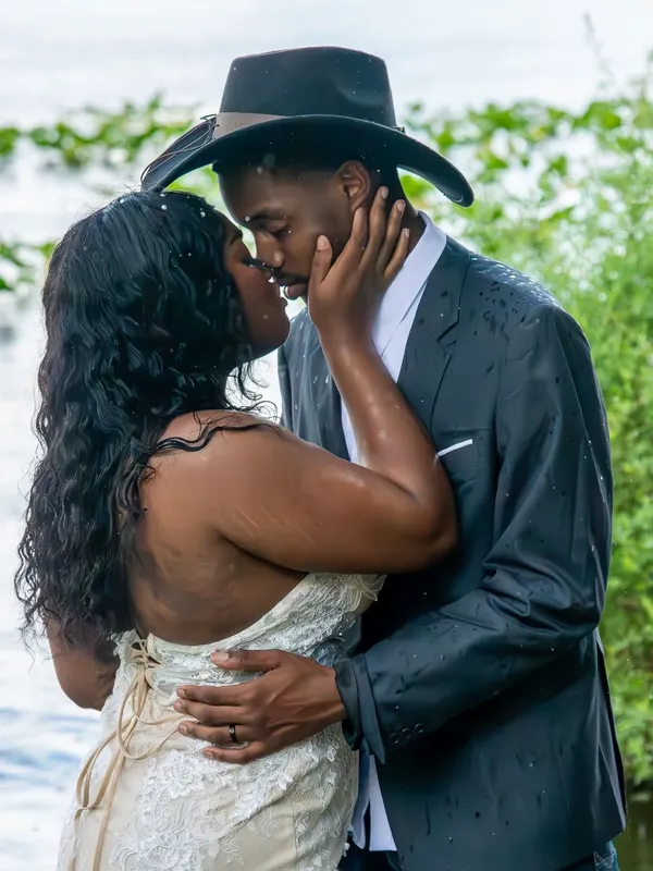 A couple in formal attire and a cowboy hat embrace tenderly near greenery and water during a light rain.